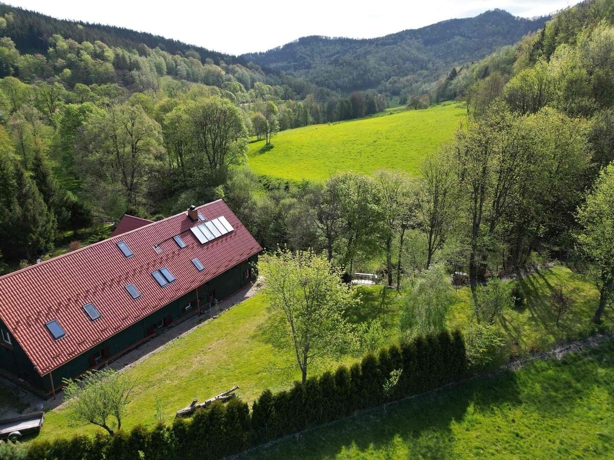 A spacious farm building with a red-tiled roof is surrounded by lush green fields and trees. Rolling hills are visible in the background, suggesting a serene natural setting. Sunlight casts a warm glow over the landscape, highlighting the vast open spaces.