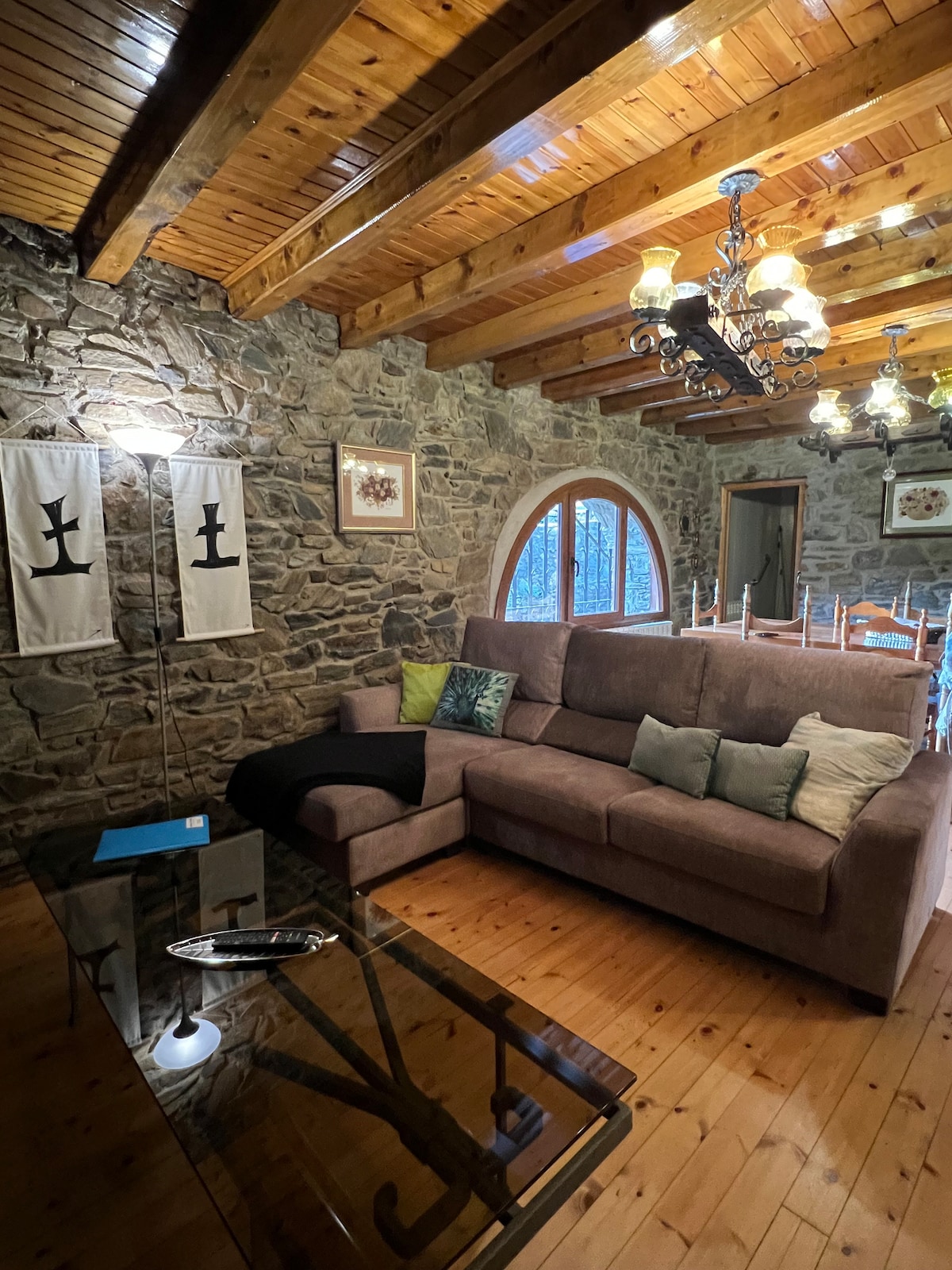 The spacious living area features a large sectional sofa adorned with decorative cushions. Exposed wooden beams stretch across the ceiling, complemented by a chandelier providing soft lighting. A glass coffee table sits in front, reflecting the warm wooden floors. Arched windows allow natural light to fill the space.