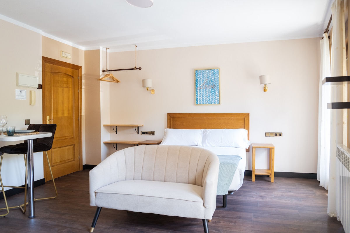 A bright and airy studio apartment is featured, with a spacious bed positioned against a light-colored wall. A comfortable couch and a small dining area with two stools are visible. Light streams in through large windows, and wood flooring adds warmth to the space.