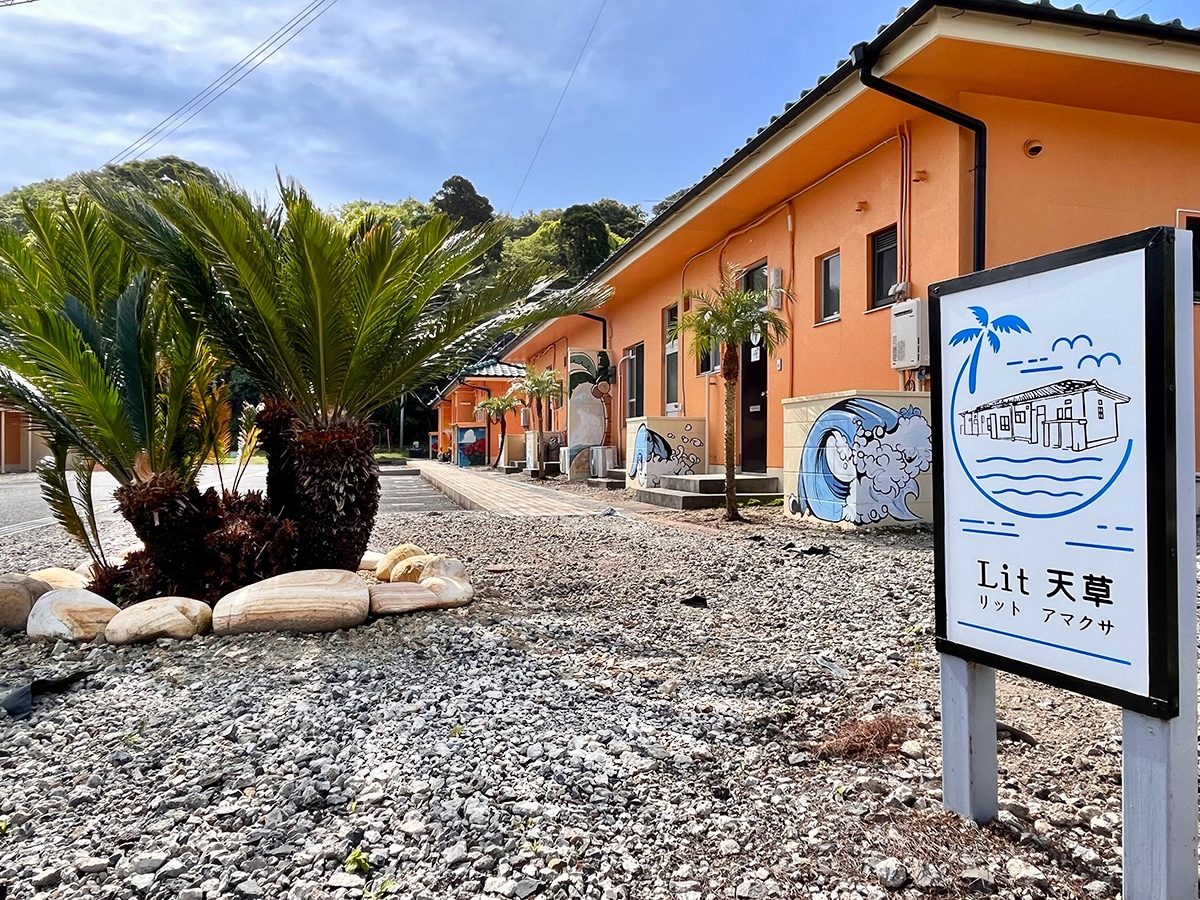 A vibrant sign displaying 'Lit 天草' stands in front of a bright orange building surrounded by gravel. Lush palm trees and ornamental plants enhance the exterior, with a mural featuring ocean waves visible on one side of the building.