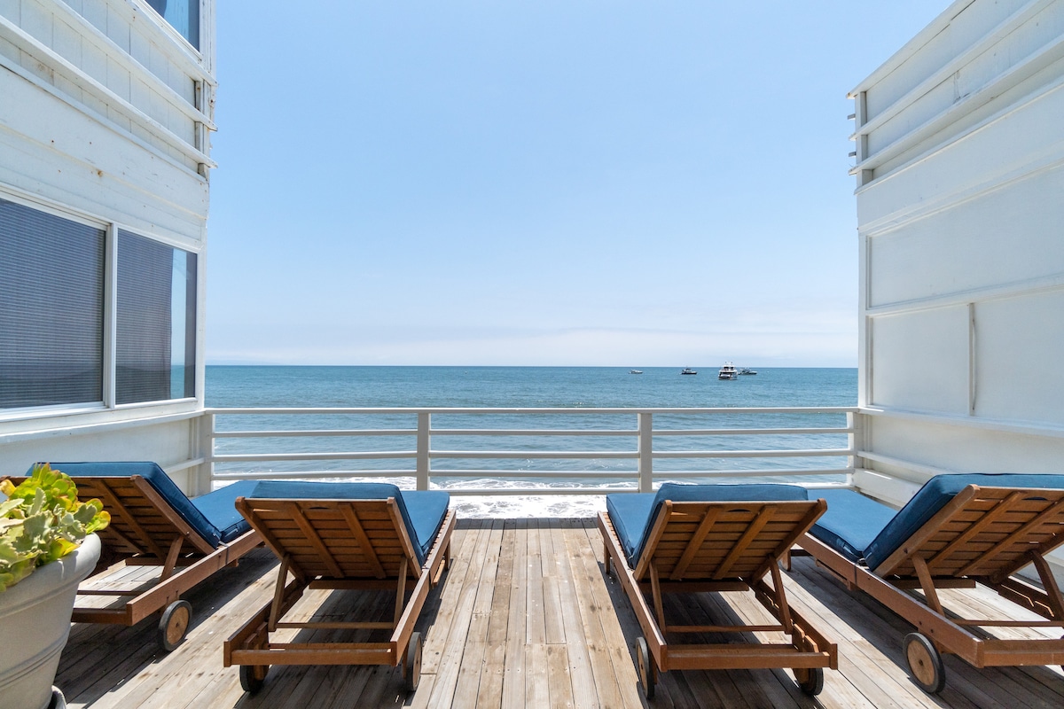 A spacious lounge area features multiple wooden loungers facing the ocean. The terrace is surrounded by a railing, offering unobstructed views of the calm water and distant boats. Bright natural light fills the scene, and a planter with greenery adds a touch of color.