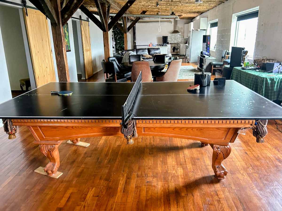 A sleek, wooden ping pong table is positioned in the foreground, showcasing its polished surface and intricately designed legs. The loft space in the background features a modern kitchen and comfortable seating areas, illuminated by natural light streaming through large windows.