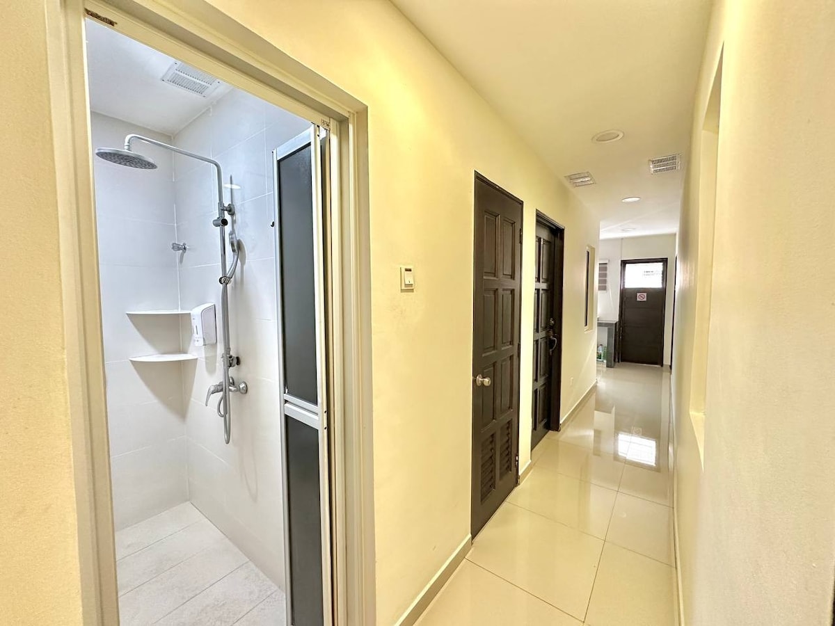 A well-lit hallway features light-colored walls and a glossy floor. Doors line both sides, leading to rooms and facilities. A wet area is visible to the left, showcasing a spacious shower with modern fixtures. Natural light illuminates the corridor, enhancing its cleanliness.