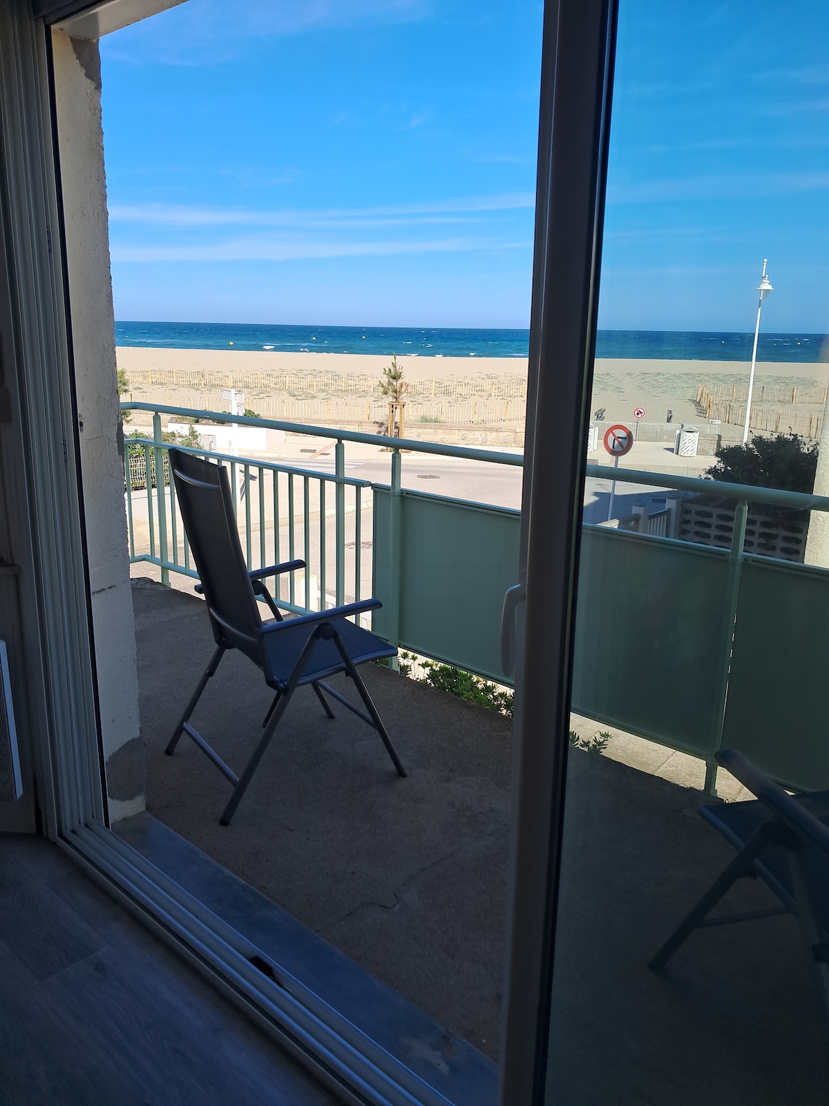 A balcony with a view of the sandy beach and the sea is depicted, featuring a single outdoor chair. The scene captures clear blue skies and gentle waves in the distance, with a railing providing a sense of openness.