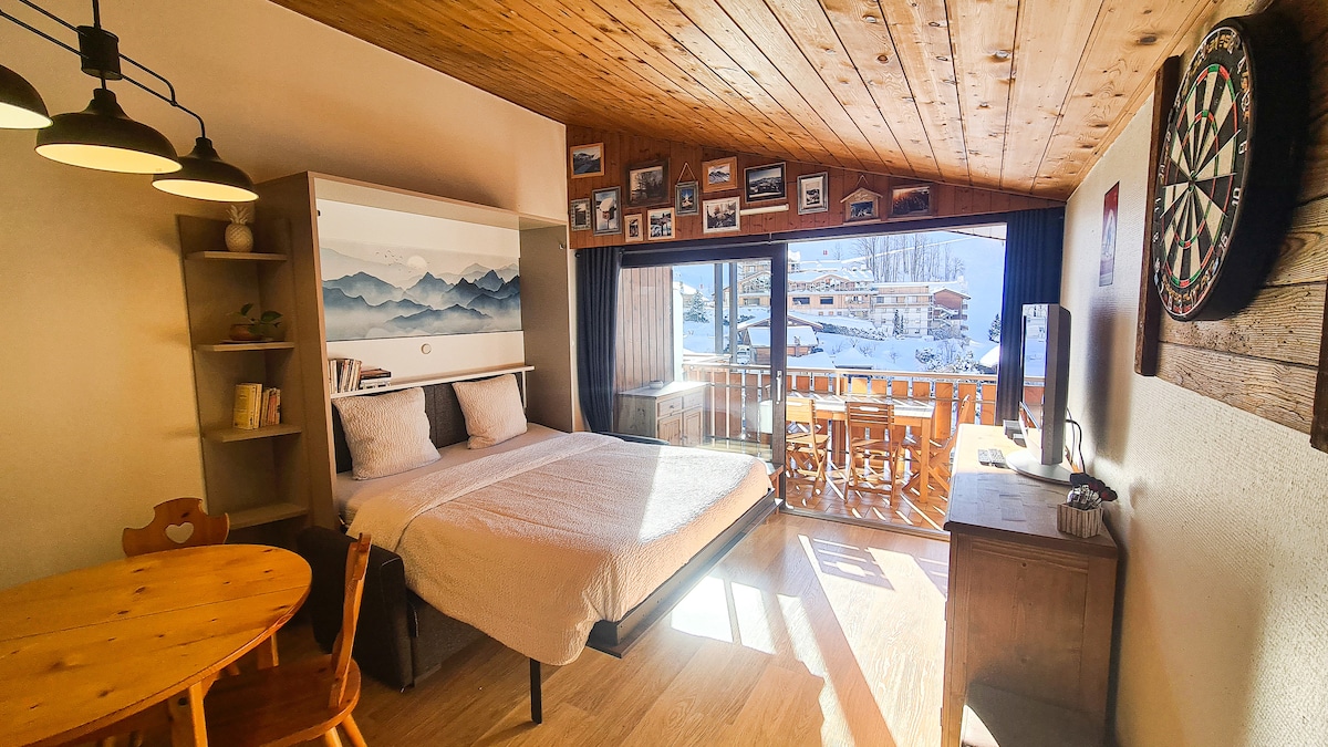A studio with wood paneling features a spacious bed positioned next to a large window, allowing natural light to fill the room. A round dining table with wooden chairs is visible, along with a TV mounted on the wall. A balcony overlooks the snowy landscape.