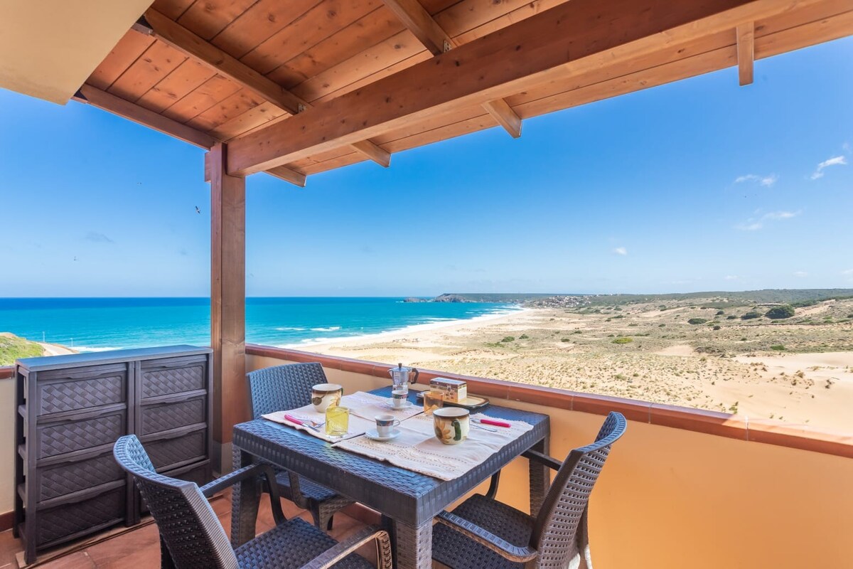 A dining area is positioned on the terrace, featuring a table set for four. The expansive view of the coastline and sand dunes is visible in the background, with the sea appearing in vibrant shades of blue under a clear sky.