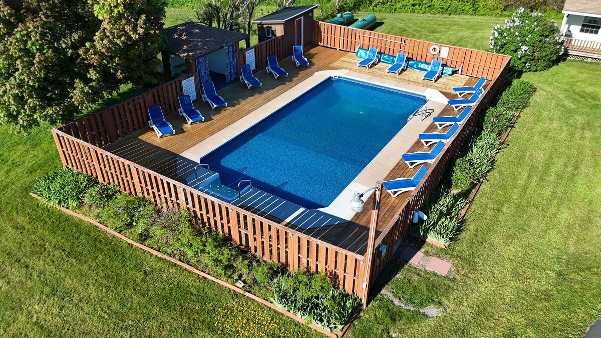 An outdoor heated pool is framed by a wooden deck, surrounded by lounge chairs and a privacy fence. Lush green landscaping borders the area, adding to the serene environment. A small shed is also visible near the pool area.