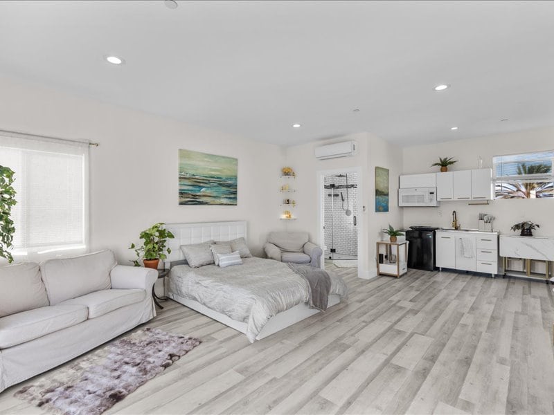 A bright studio space features a queen bed with soft bedding and a cozy sofa nearby. The well-equipped kitchen, complete with utensils and a fridge, is visible. Natural light illuminates the room through large windows adorned with light curtains, while potted plants add a touch of greenery.