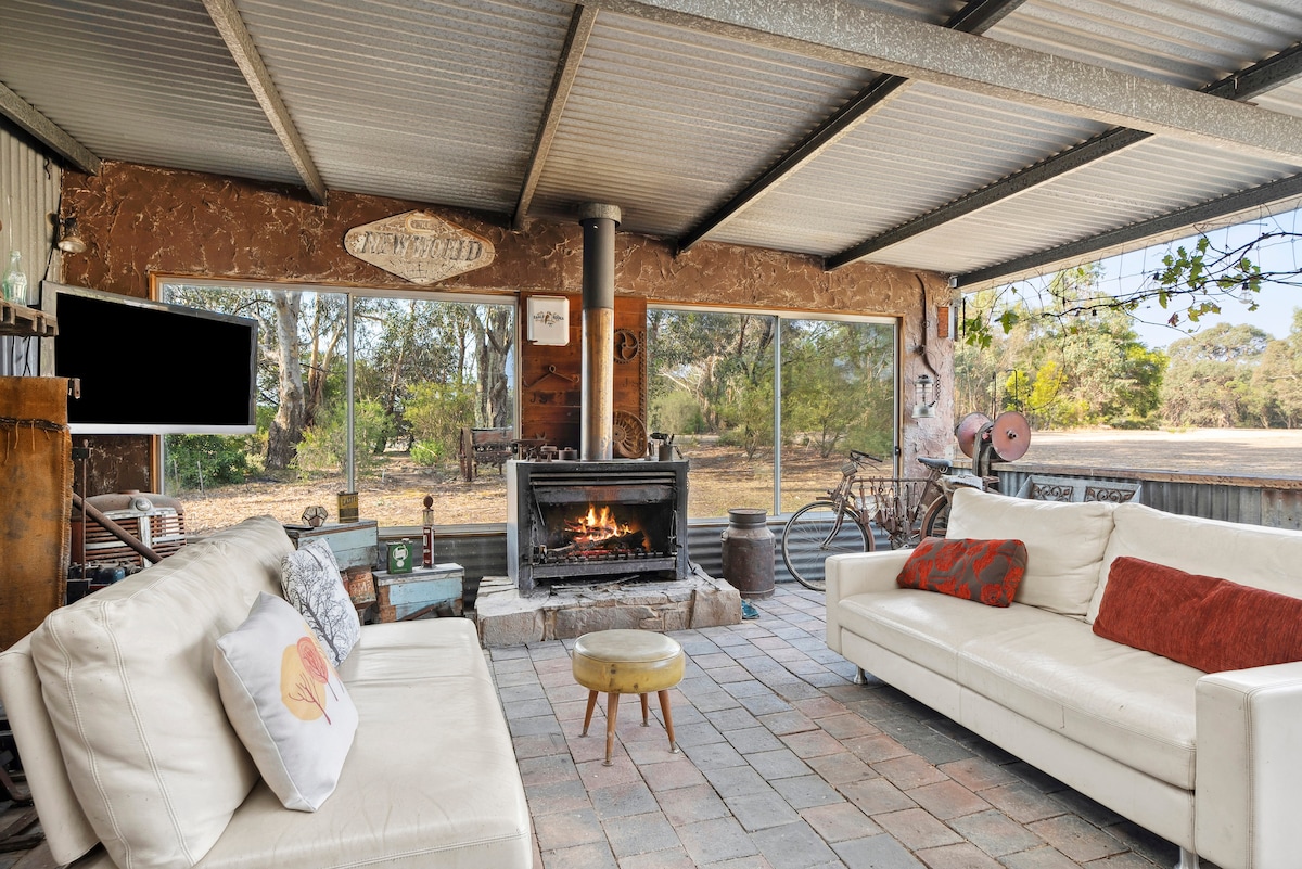 An inviting outdoor entertainment area features a cozy seating arrangement with white sofas, arranged around a large stone fireplace. Large windows allow natural light to fill the space, while views of the surrounding nature create a peaceful atmosphere.