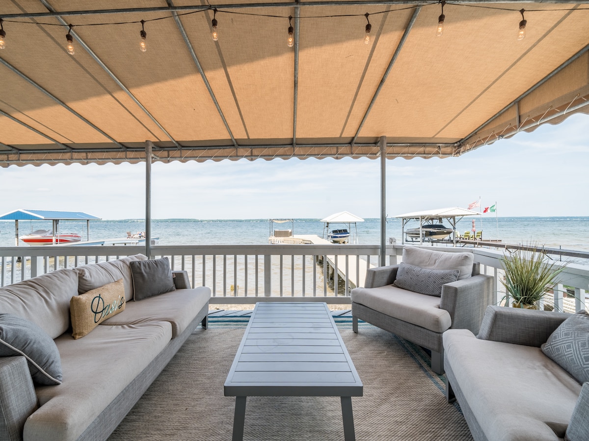 A covered deck provides comfortable seating arrangements with soft gray sofas and a coffee table. The area overlooks Seneca Lake, featuring a clear view of the water and dock. Soft lighting is strung across the awning, enhancing the tranquil outdoor space.