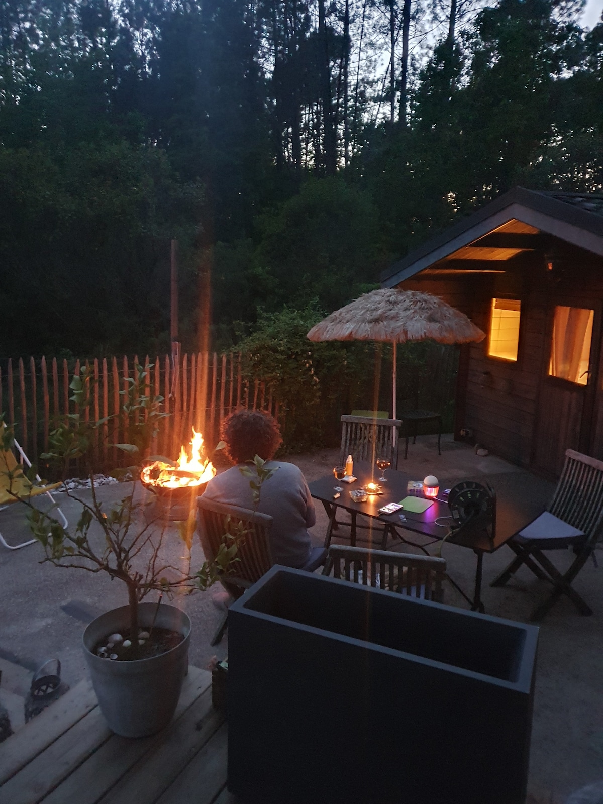 An outdoor seating area is featured with wooden chairs surrounding a low table, illuminated by candlelight. A small fire burns nearby, contributing to a cozy atmosphere. A thatched umbrella provides shade, and the guesthouse is visible in the background, softly lit through its window.