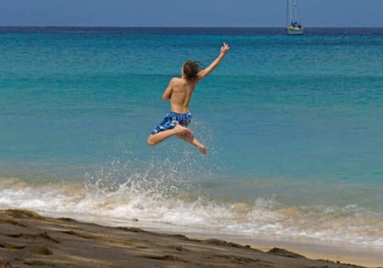 A child is captured mid-air, joyfully leaping into the clear, turquoise ocean waves. The sandy shore is visible, with gentle waves splashing at the child's feet. A sailboat can be seen in the distance against a serene sky.