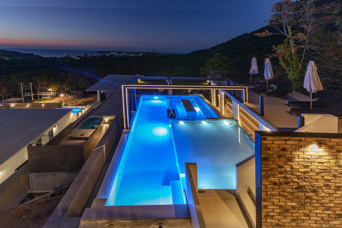 An illuminated swimming pool is showcased at night, with vibrant blue lights reflecting off the water's surface. Surrounding structures feature modern architecture, while umbrellas stand elegantly on the edges. The distant mountains create a serene backdrop, enhancing the peaceful atmosphere.