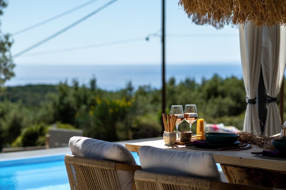 An outdoor dining area is set with glasses and utensils on a wooden table. Soft sunlight highlights the inviting pool and the sea view in the background, while surrounding greenery adds a natural touch to the serene setting.