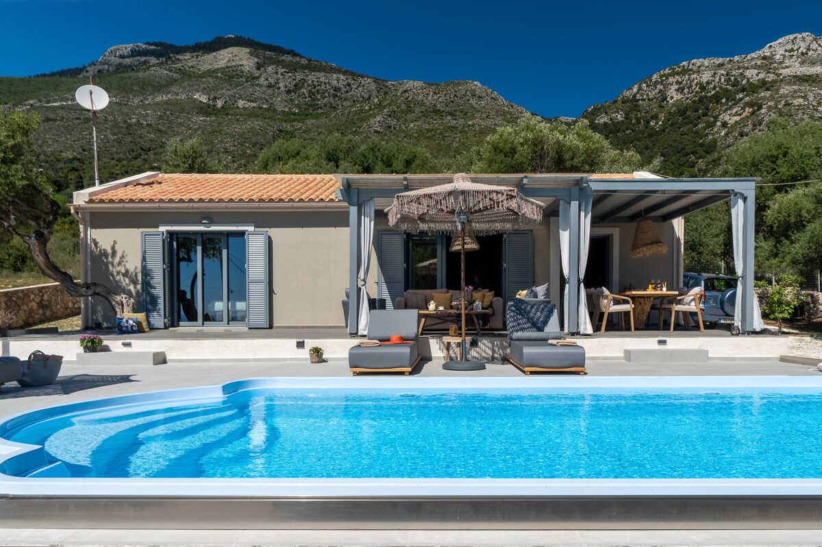 A serene outdoor area features a sparkling pool surrounded by sun loungers, with a shaded seating arrangement under a fabric canopy. The house, with sliding glass doors, opens to the poolside, set against a backdrop of lush greenery and hills.