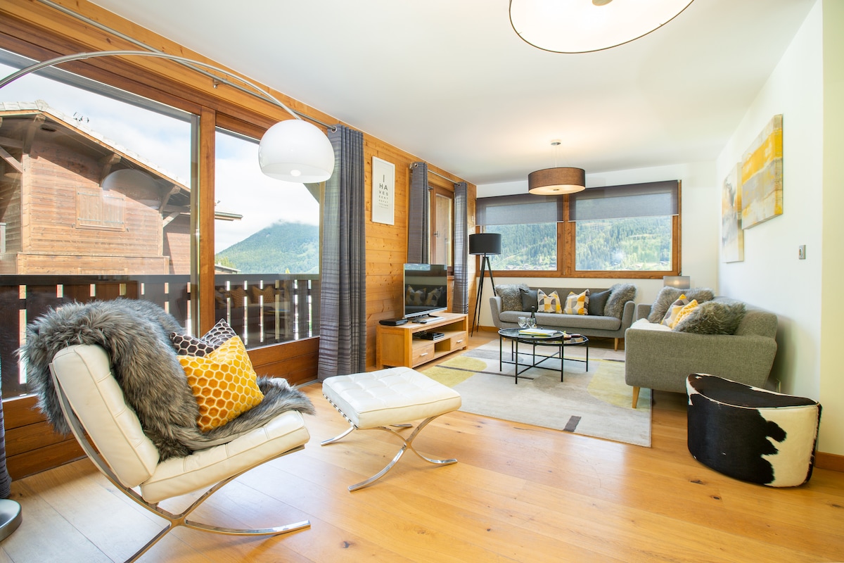 The spacious lounge area features large floor-to-ceiling windows offering views of the surrounding mountains. Comfortable seating includes a plush sofa and a contemporary lounge chair. A flat-screen TV is positioned on a wooden console, while modern décor enhances the inviting ambiance.