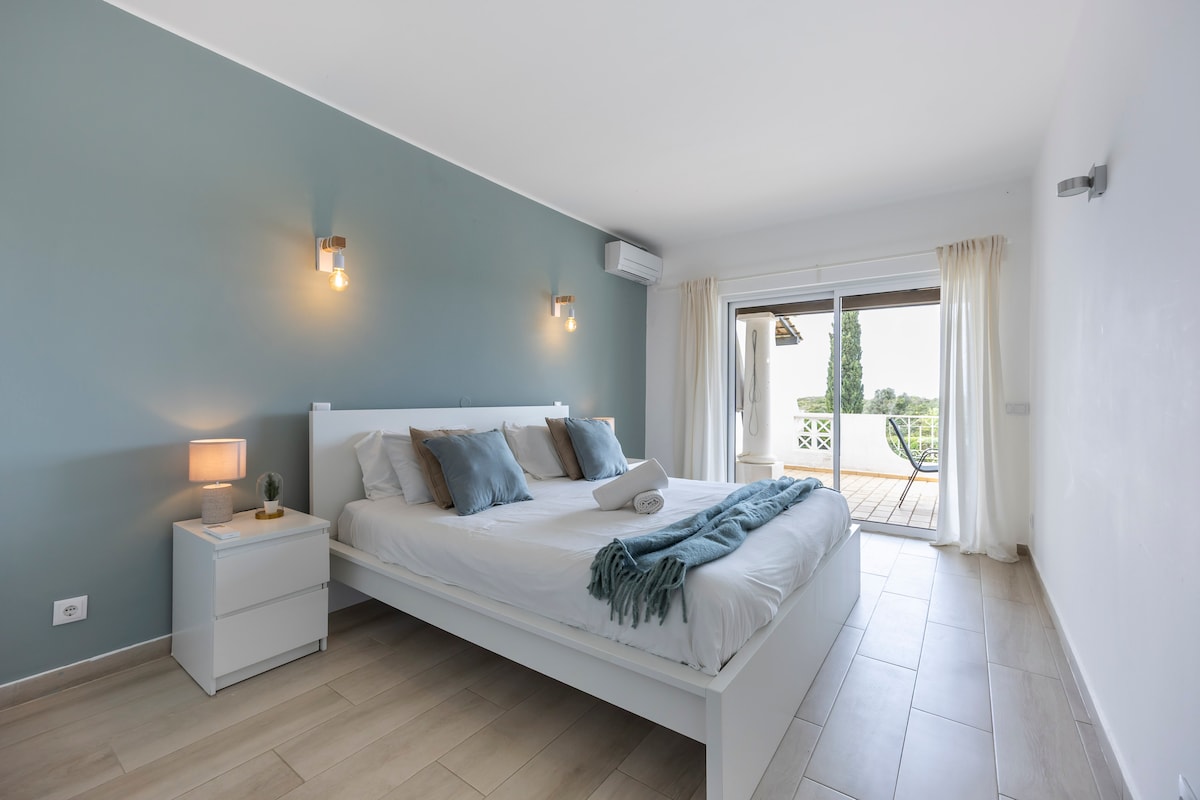 A bright bedroom features a spacious bed dressed in crisp white linens, complemented by decorative pillows in soft tones. A small bedside table holds a lamp, and large glass doors open to a balcony with outdoor seating, filling the room with natural light.