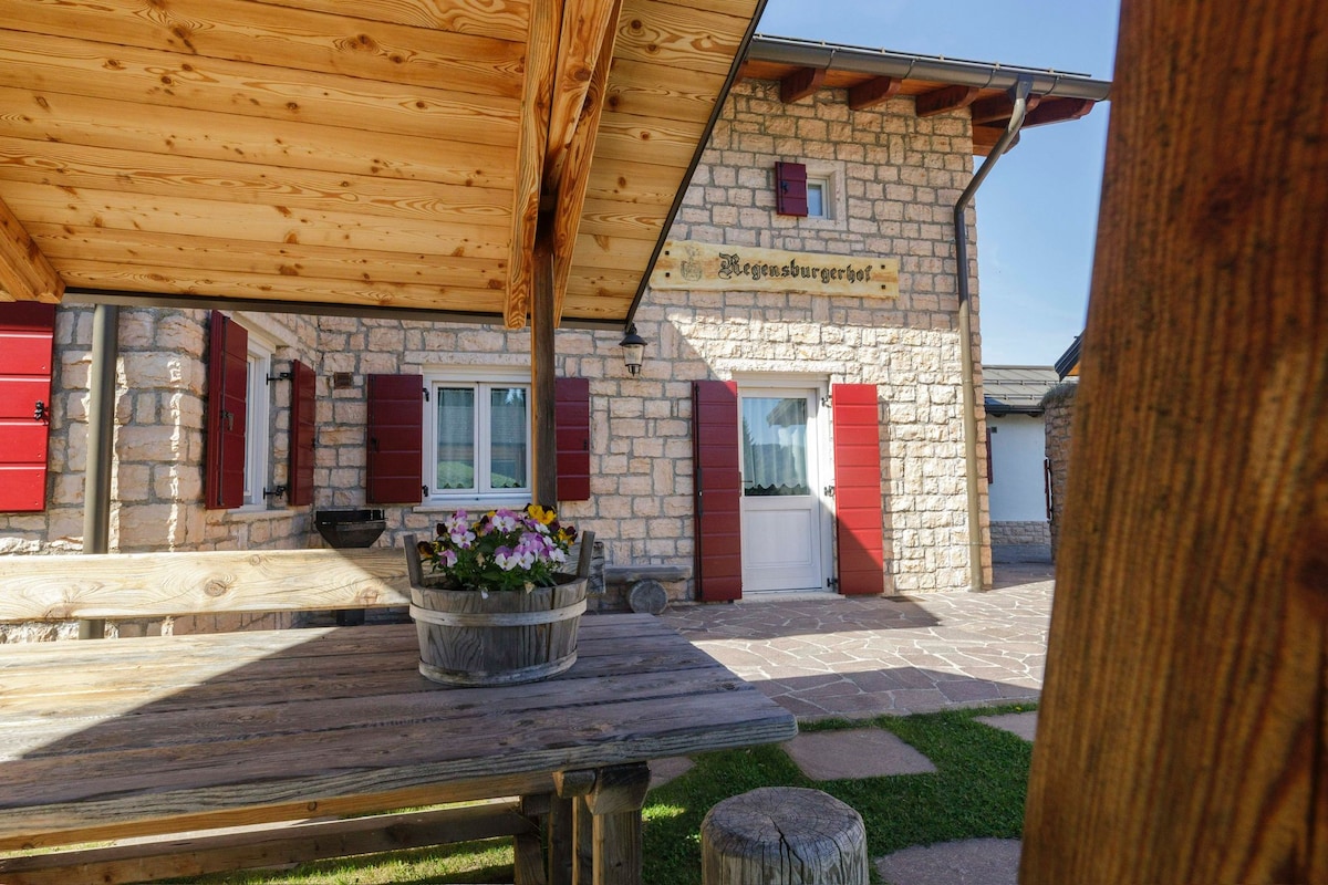 A rustic wooden table and benches are situated beneath a charming awning. The stone facade of the property features red shutters and a welcoming entrance door. A wooden planter filled with colorful flowers adds a touch of nature.