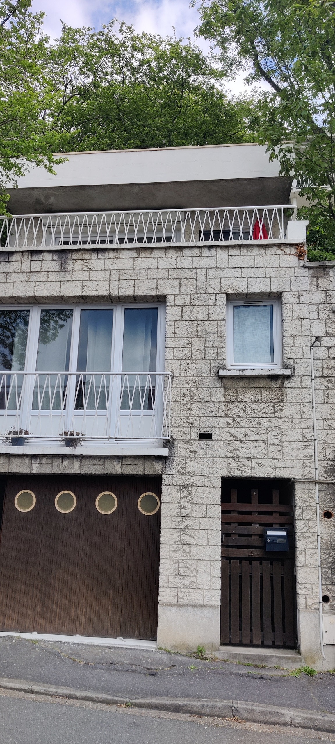 A two-story structure is displayed with a stone façade and modern balcony railing. Large windows allow ample natural light, and the entrance features a wooden door with round decorative windows above. Green foliage from nearby trees adds a touch of nature surrounding the building.