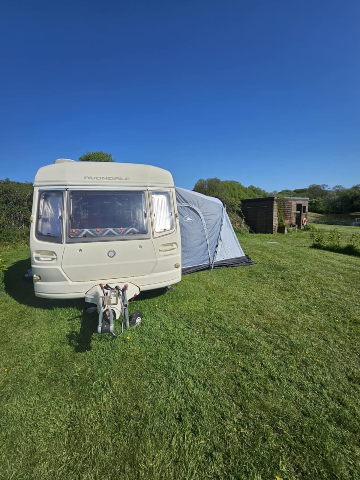 A touring caravan is parked on a grassy area, complemented by an attached awning. The clear blue sky above enhances the serene atmosphere, while trees border the campsite. A storage shed is visible in the background, providing additional utility to the outdoor setting.