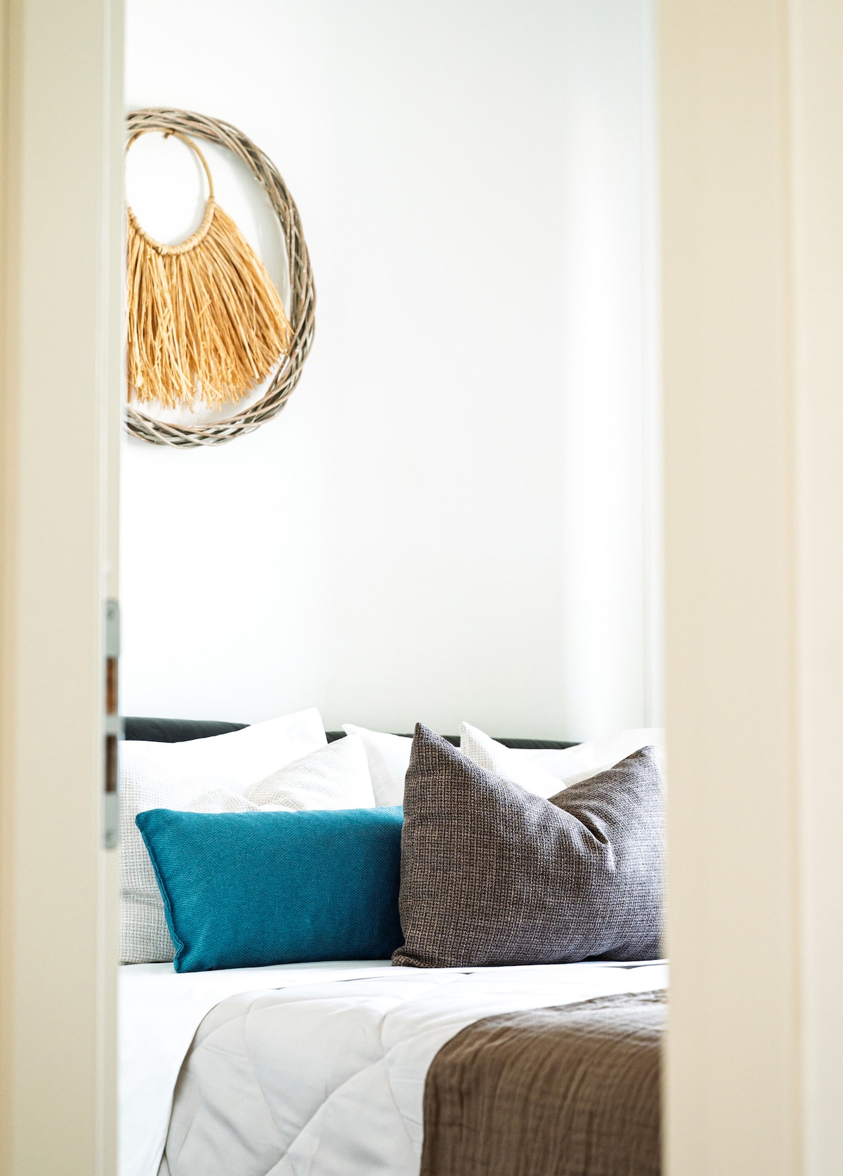 A glimpse into a serene sleeping area features a bed adorned with a mix of textured pillows in shades of turquoise and gray. A decorative wall piece made of natural fibers hangs above, complementing the neutral color scheme. Soft bedding creates an inviting look.