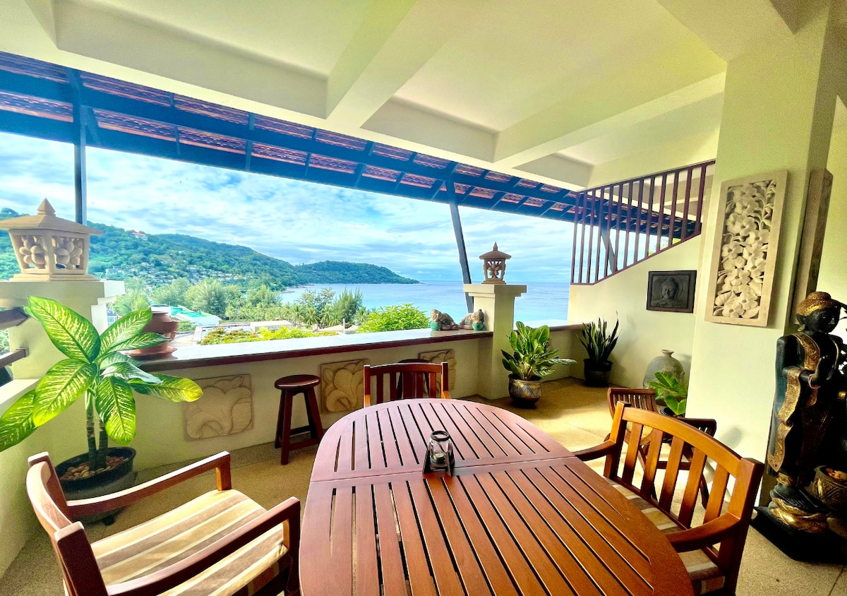 An inviting terrace area features a wooden dining table surrounded by four chairs. Lush green plants are positioned around the space, while the stunning view of the ocean is framed by the terrace's overhang, enhancing the calming atmosphere.