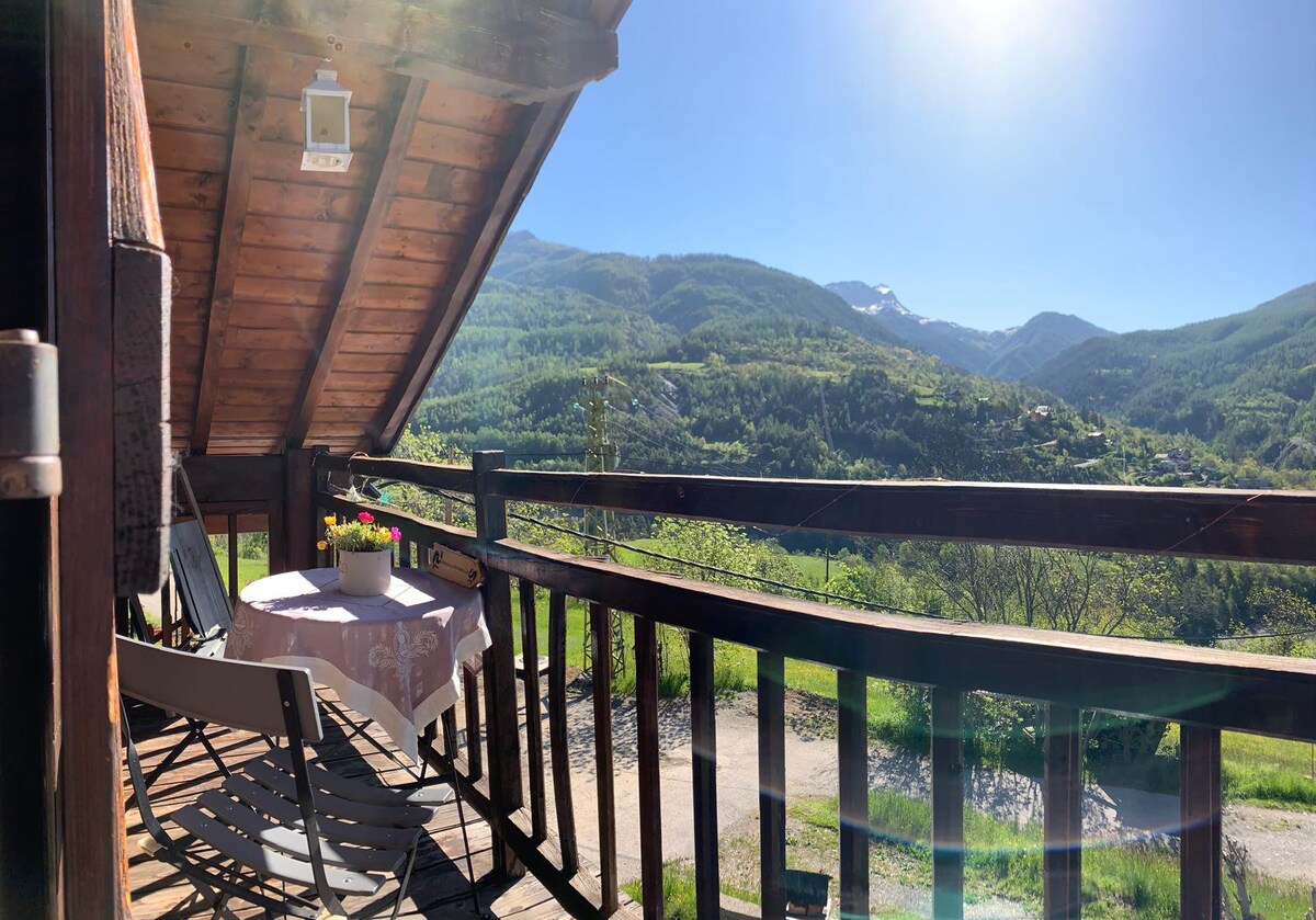 A covered terrace with a round table and chairs offers scenic views of the surrounding mountains and valleys. Sunlight bathes the area, highlighting the greenery and distant peaks, creating a serene outdoor retreat.