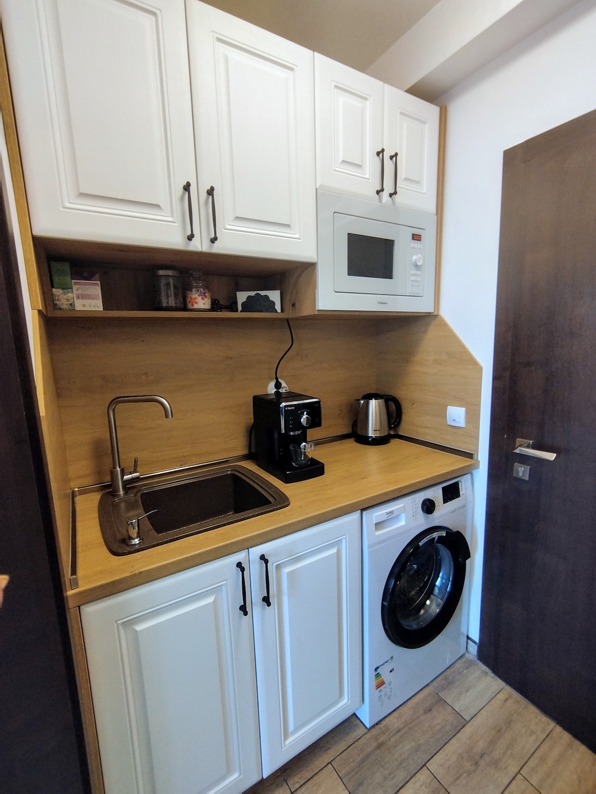 The compact kitchen features white cabinetry and a wooden countertop. A coffee maker, microwave, and kettle are visible alongside a sink and a washing machine. Essential appliances are arranged for convenience, creating an efficient space for meal preparation and laundry.