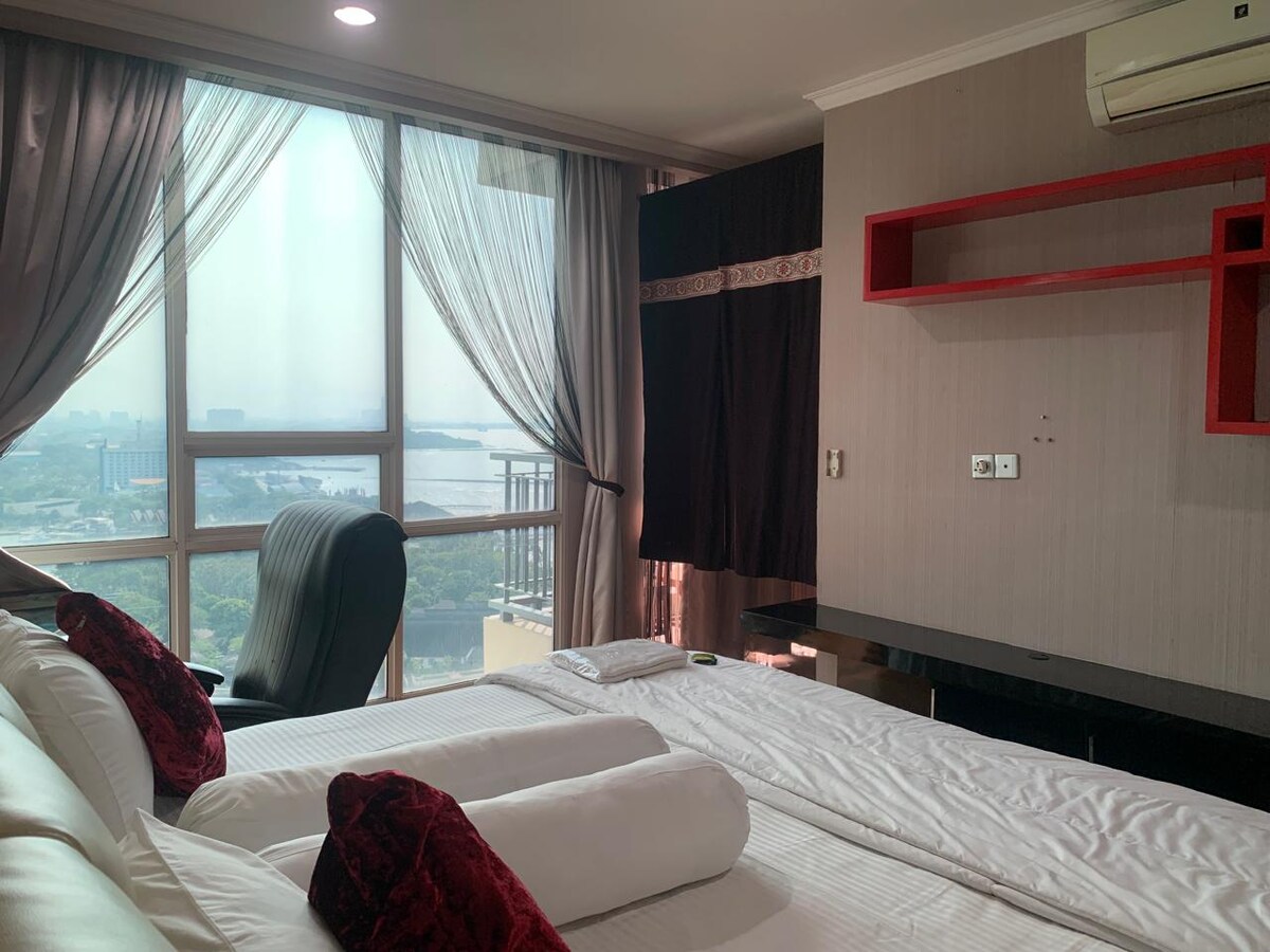 A well-lit bedroom features a spacious bed with two rolled white towels and burgundy accent pillows. A large window offers views of the surrounding area, while sheer curtains allow natural light to filter in. A black office chair is positioned near a minimalist desk.