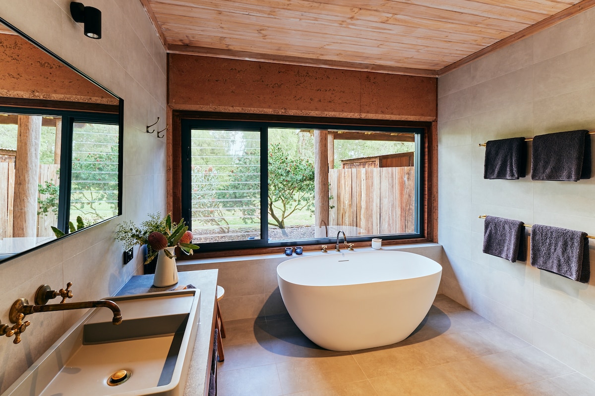 A modern bathroom features a freestanding stone bath positioned next to large windows that provide views of the outdoor garden. A sleek vanity with a stylish sink is complemented by a mirror above. Towels hang neatly on the wall, enhancing the overall functionality.