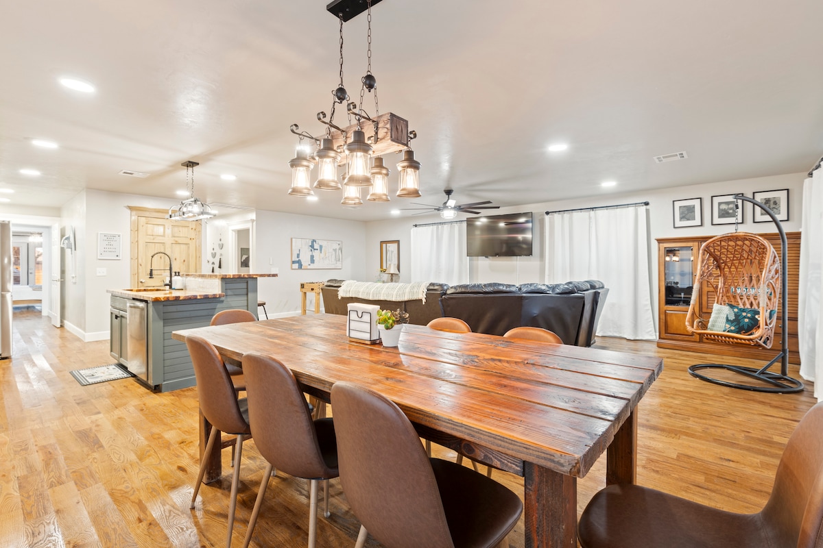 A spacious dining area is featured with a rustic wooden table and brown chairs, illuminated by a distinctive pendant light. In the background, a cozy living area is visible, showcasing a large sectional sofa and a wall-mounted television, creating an inviting communal space.