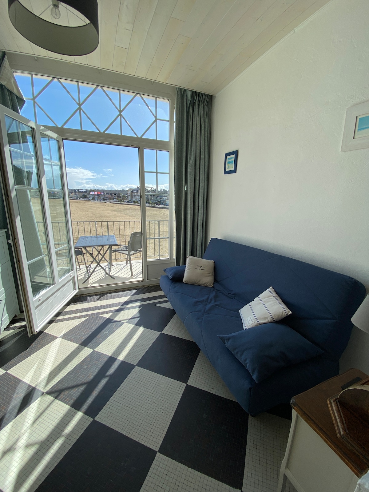A bright living area features a blue futon and a small side table with two chairs. Large glass doors open to a balcony, allowing natural light to fill the space. The checkerboard floor adds a classic touch to the room.