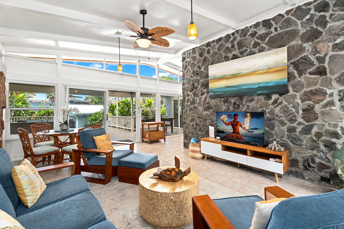 A spacious living room features a comfortable seating arrangement with blue sofas and tropical decor. A natural lava stone wall serves as a focal point, while a large TV is positioned for entertainment. Abundant natural light enters through multiple windows, enhancing the inviting space.