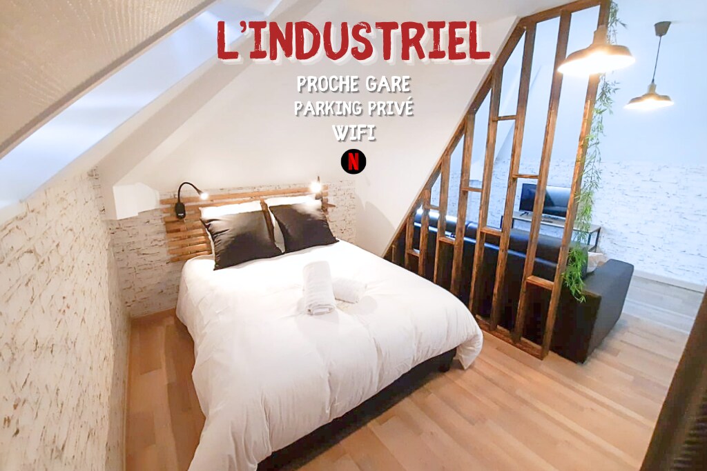 A cozy bedroom features a spacious bed with white bedding, accompanied by a rustic wooden headboard. A side table with a wall-mounted lamp is present. The area is illuminated by overhead lighting, with a comfortable couch visible in the background.