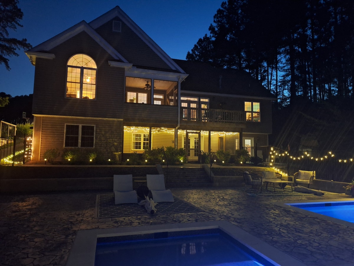 The exterior of the home is illuminated by soft lights, creating a warm atmosphere at night. The backyard features two swimming pools adjacent to a concrete deck with various seating options. Tall trees provide a serene backdrop to the scene.