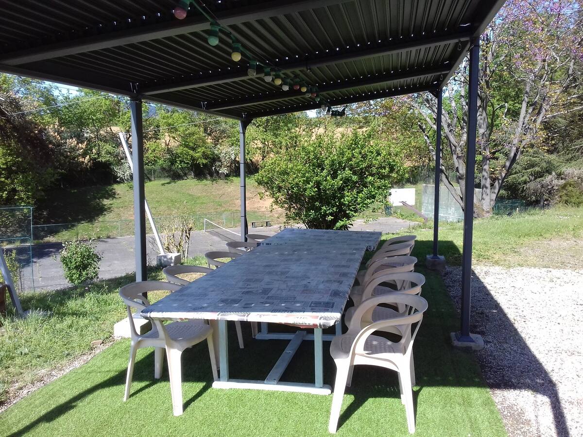 An outdoor dining area is set under a covered terrace, featuring a long table surrounded by multiple light-colored chairs. The space is fitted with string lights above, and greenery can be seen in the background, providing a serene environment.