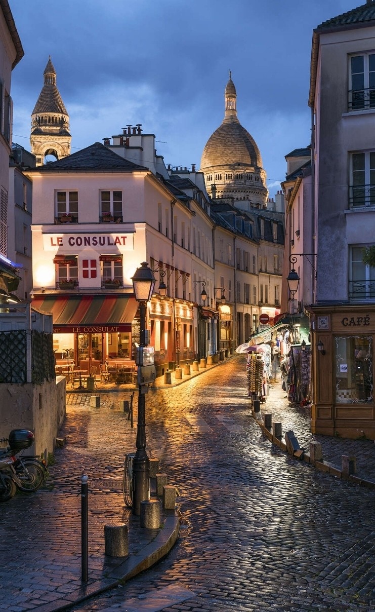 A charming street in Montmartre is shown, lined with illuminated restaurants and shops. The cobblestone road glistens from recent rain, and the domes of Sacré-Cœur rise majestically in the background, blending into the evening sky. Soft lighting adds a welcoming glow to the scene.