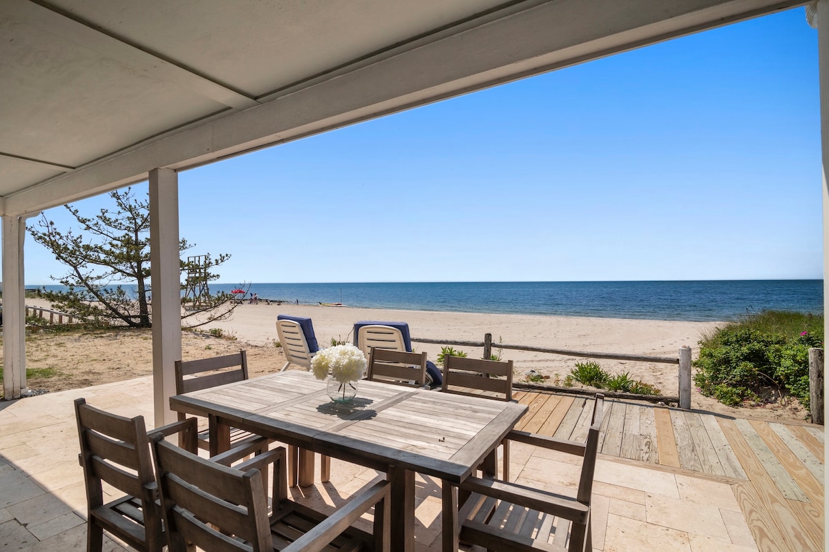 An outdoor dining area features a wooden table surrounded by chairs, facing the beach. The view overlooks sandy shores and the ocean under a clear blue sky. Green plants and a tree are positioned nearby, adding to the natural surroundings.