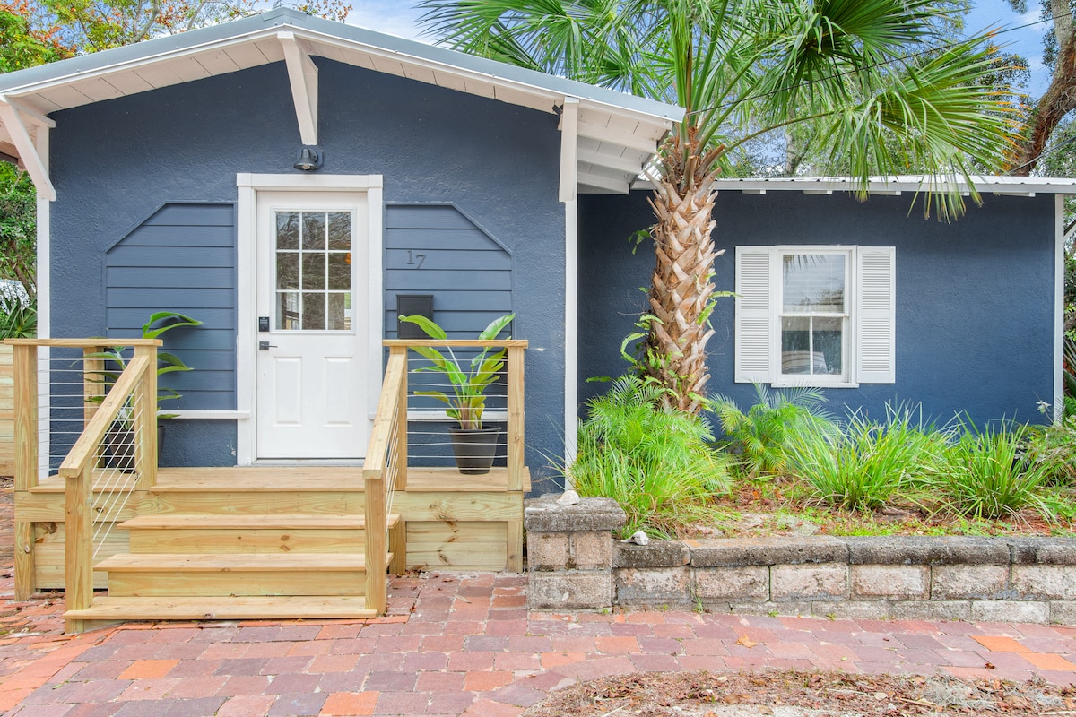 The exterior of the cottage is showcased, featuring a deep blue facade and a welcoming front door flanked by plants. A wooden deck with steps leads to the entrance, and a palm tree along with other greenery enhances the inviting appearance.
