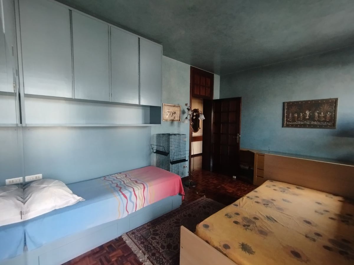 The room features two beds, each dressed in different colorful linens. A patterned rug lies on the wooden floor, and a small cage is positioned in one corner. Above the beds, built-in cabinetry offers additional storage space. Natural light enters through a nearby window.