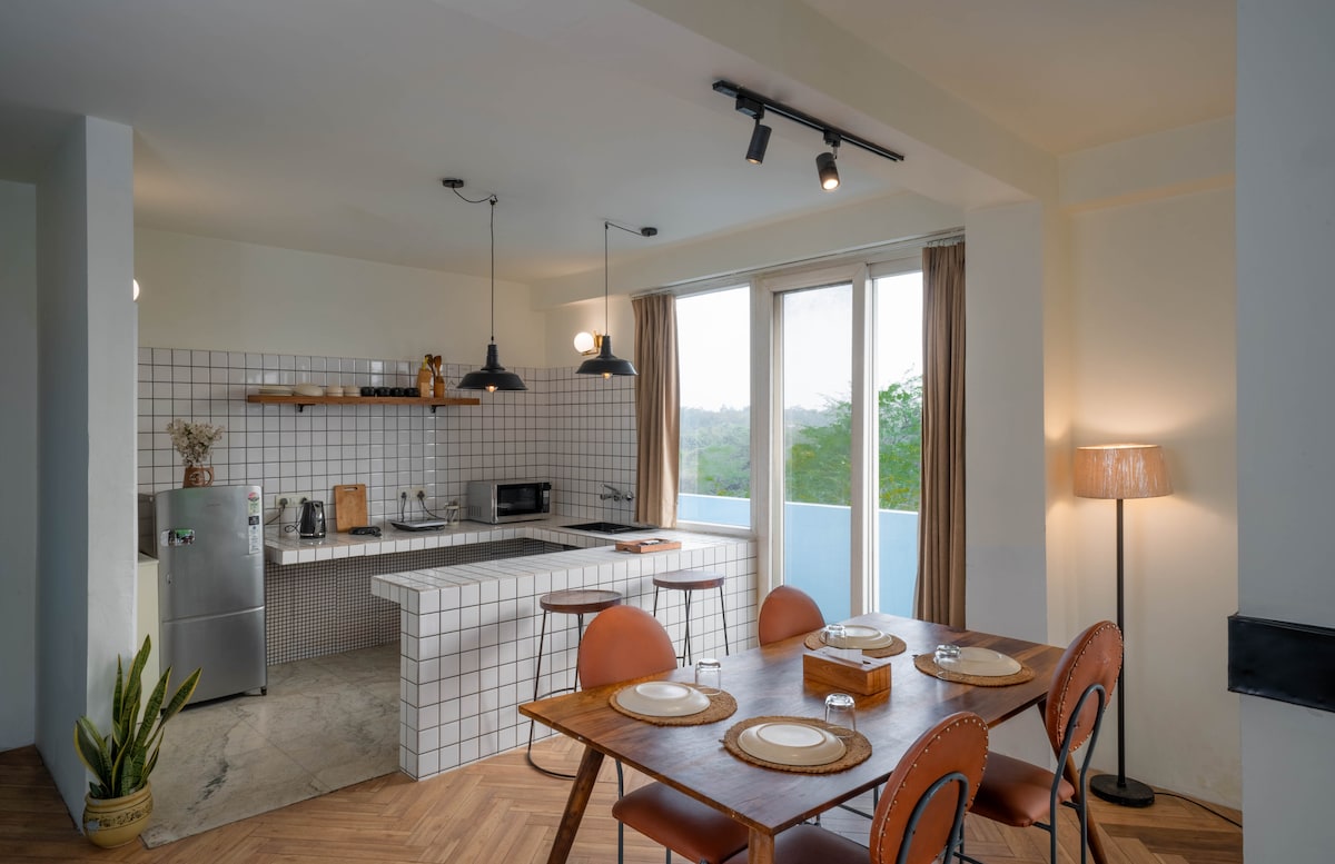 A minimalist kitchen and dining area features a simple wooden dining table surrounded by leather chairs. The space includes a modern kitchenette with white tiled backsplash, a stainless steel refrigerator, and a large window offering a view of greenery. Soft lighting enhances the airy atmosphere.