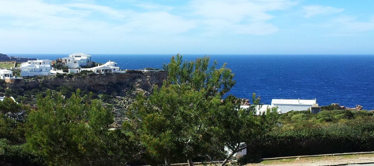 A scenic view of the coastline is captured, featuring gentle waves lapping against rocky shores. White buildings are visible on the cliffs, surrounded by lush greenery. The sky is clear with scattered clouds, enhancing the overall peacefulness of the natural setting.
