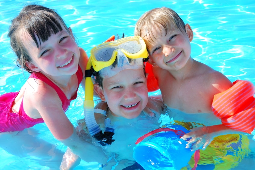 Two children are enjoying playtime in a bright blue swimming pool. One child wears a yellow snorkel mask and flippers, while the other child dons colorful arm floats. Vibrant pool toys are scattered nearby, adding a lively touch to the scene.