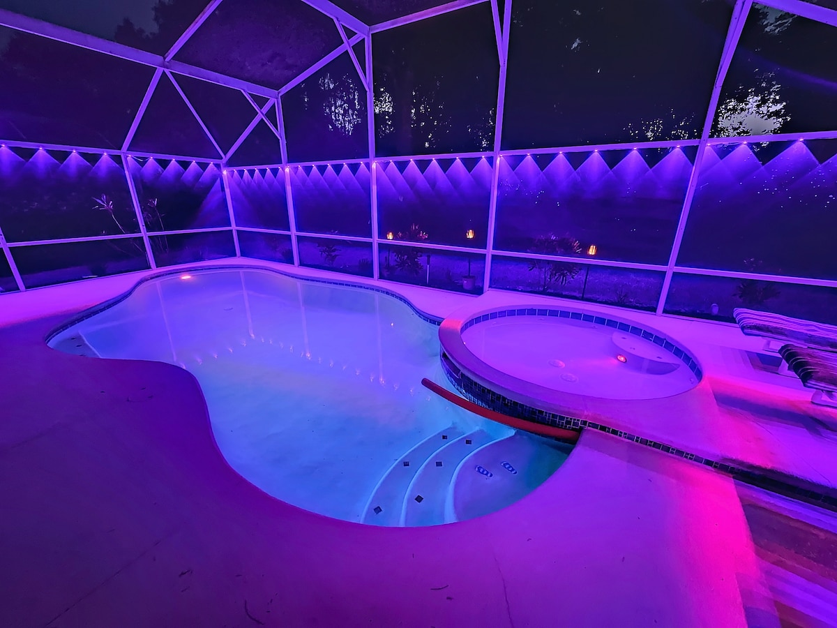 A private pool area is showcased, featuring a spacious pool with an integrated spa. The space is illuminated with soft purple lighting, enhancing the tranquil atmosphere. The screened enclosure provides protection while allowing views of the surrounding greenery.