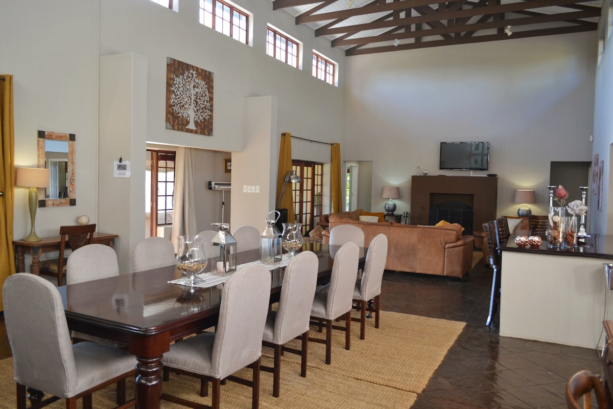 A spacious dining area features a large wooden table surrounded by upholstered chairs. Natural light enters through multiple windows, highlighting the high ceilings with wooden beams. A comfortable seating area is visible in the background, equipped with a flat screen television and a cozy fireplace.