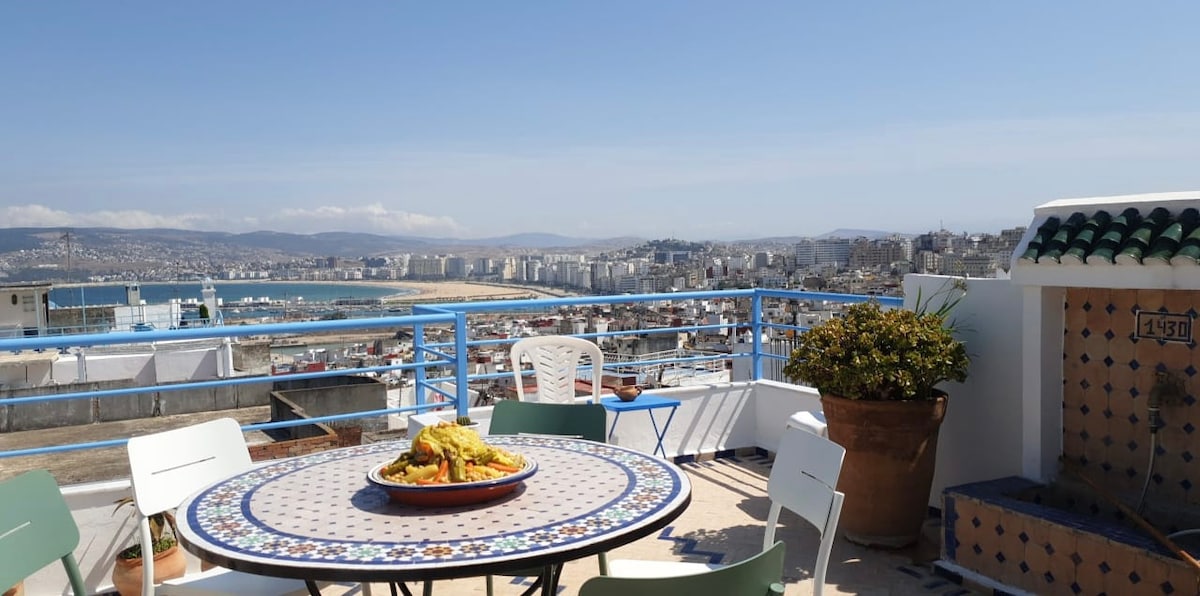A rooftop terrace offers panoramic views of the bay and city. A colorful tiled table is set with a traditional dish, surrounded by white chairs. The blue railings delineate the space, enhancing the outdoor experience against a clear sky.