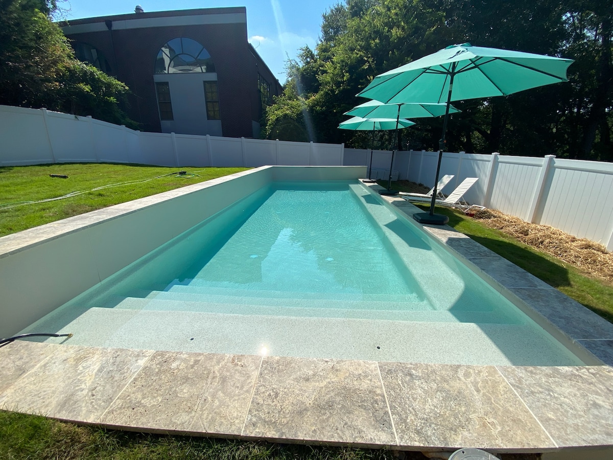 A refreshing pool is set in a spacious yard, surrounded by a neatly trimmed lawn and white fencing. Light blue umbrellas offer shade near the poolside, and the water appears clear and inviting, reflecting sunlight on the surface.
