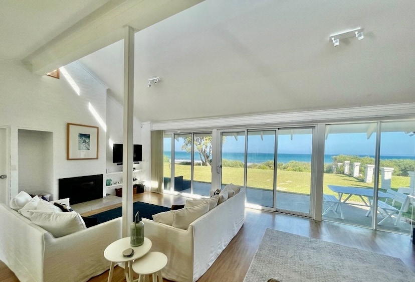 An open living area is visible, featuring a light-colored sofa and an accent chair, positioned around a central coffee table. Large glass doors frame a view of the ocean, allowing natural light to fill the space. A modern television is mounted on the wall opposite the seating.