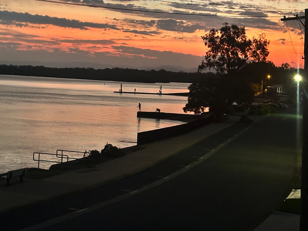 The image captures a serene riverside scene at dusk, with a vibrant sunset reflecting on calm waters. Silhouettes of individuals fishing from a pontoon are visible, alongside gentle waves lapping against the shore. Street lights cast a soft glow on the nearby pathway.