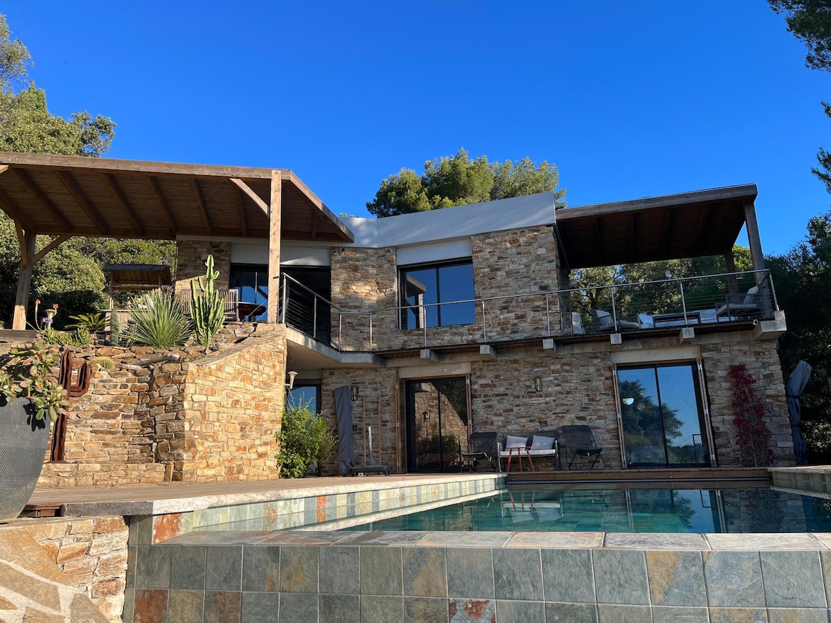 A modern villa constructed with natural stone is showcased, featuring large glass doors that open to a private pool area. A spacious terrace with a wooden roof provides shade, while greenery surrounds the property, complementing the blue sky above.
